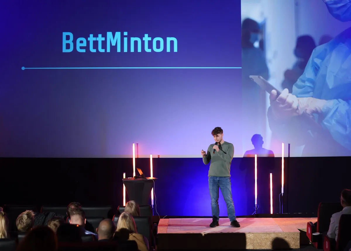 Leon Schönberg pitcht BettMinton auf der Bühne, hinter ihm der Name auf der Leinwand